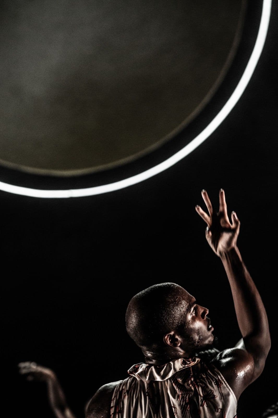 Close-up of dancer Donovan Reed reaching one arm upward toward a large glowing illuminated ring against a dark backdrop.