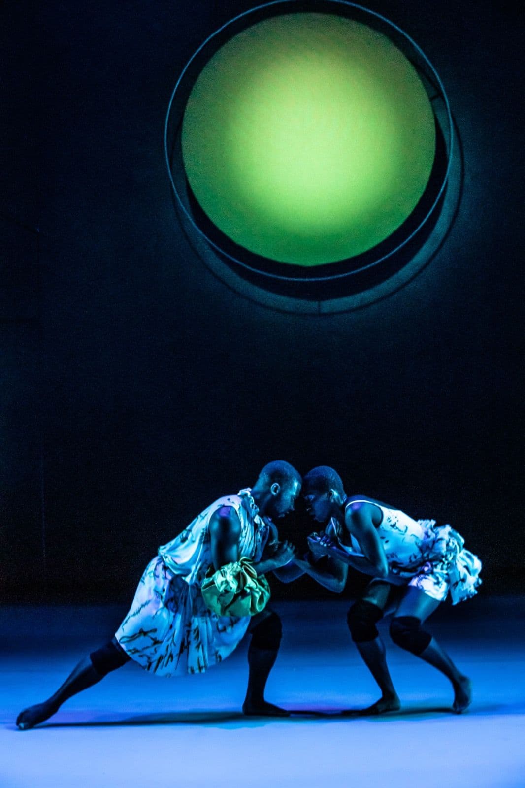 Two dancers in patterned dresses leaning toward each other head-to-head under a large glowing green orb light.