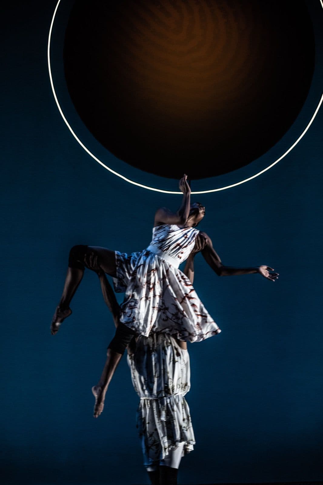 A dancer lifted high into the air by another performer beneath a large glowing ring backdrop.