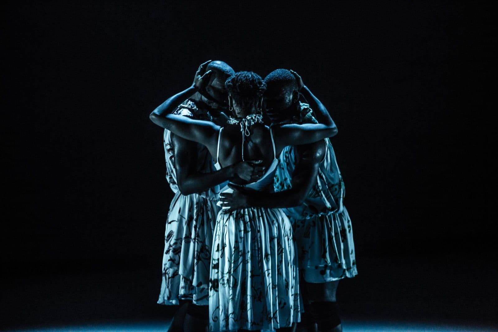Three dancers in patterned dresses standing in a close, supportive embrace under cool blue stage lighting.