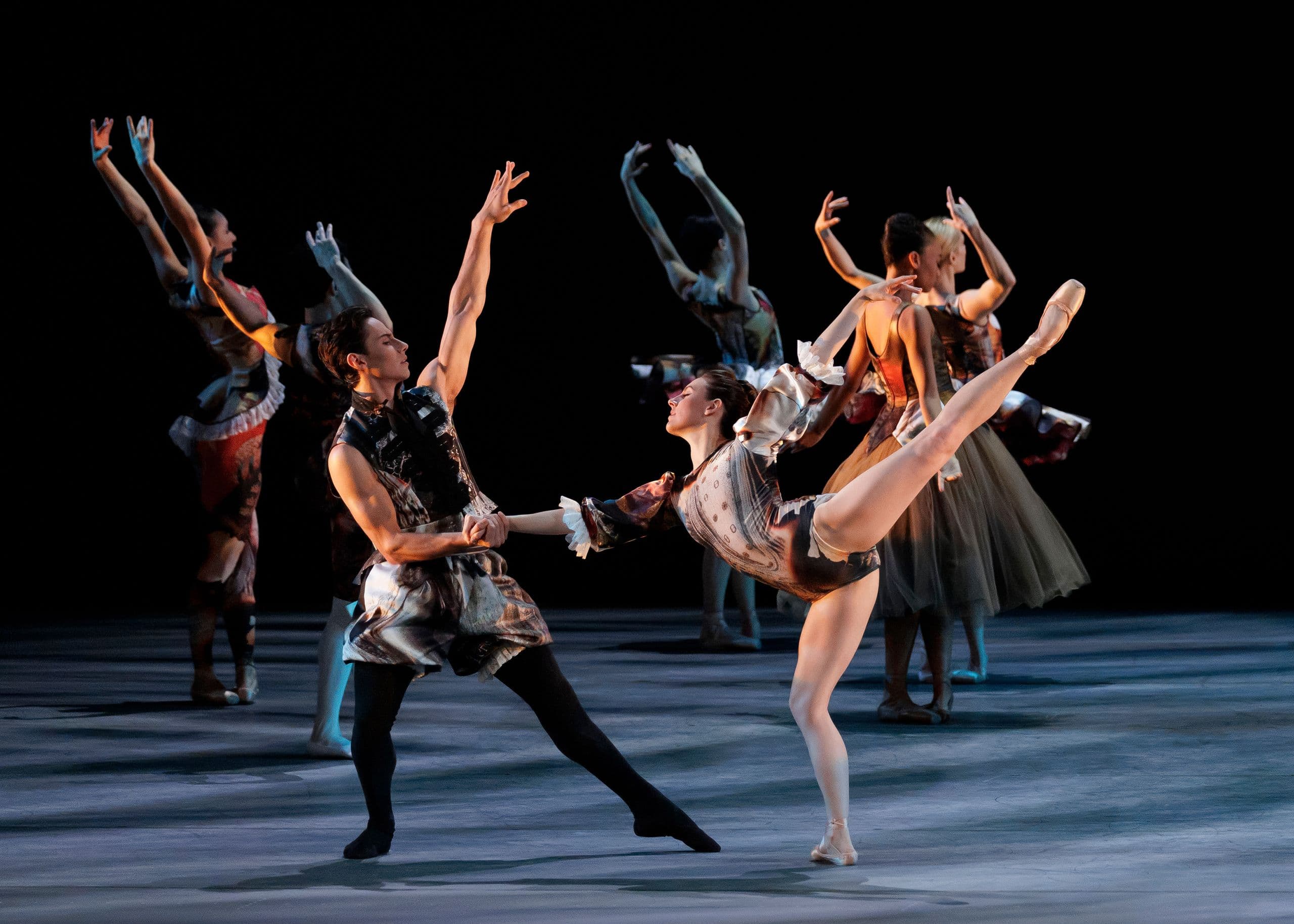 A ballet partner lifts a dancer's leg high into an arabesque as she leans back, hand in hand with him on a dimly lit stage, ensemble dancers raising their arms in the background.