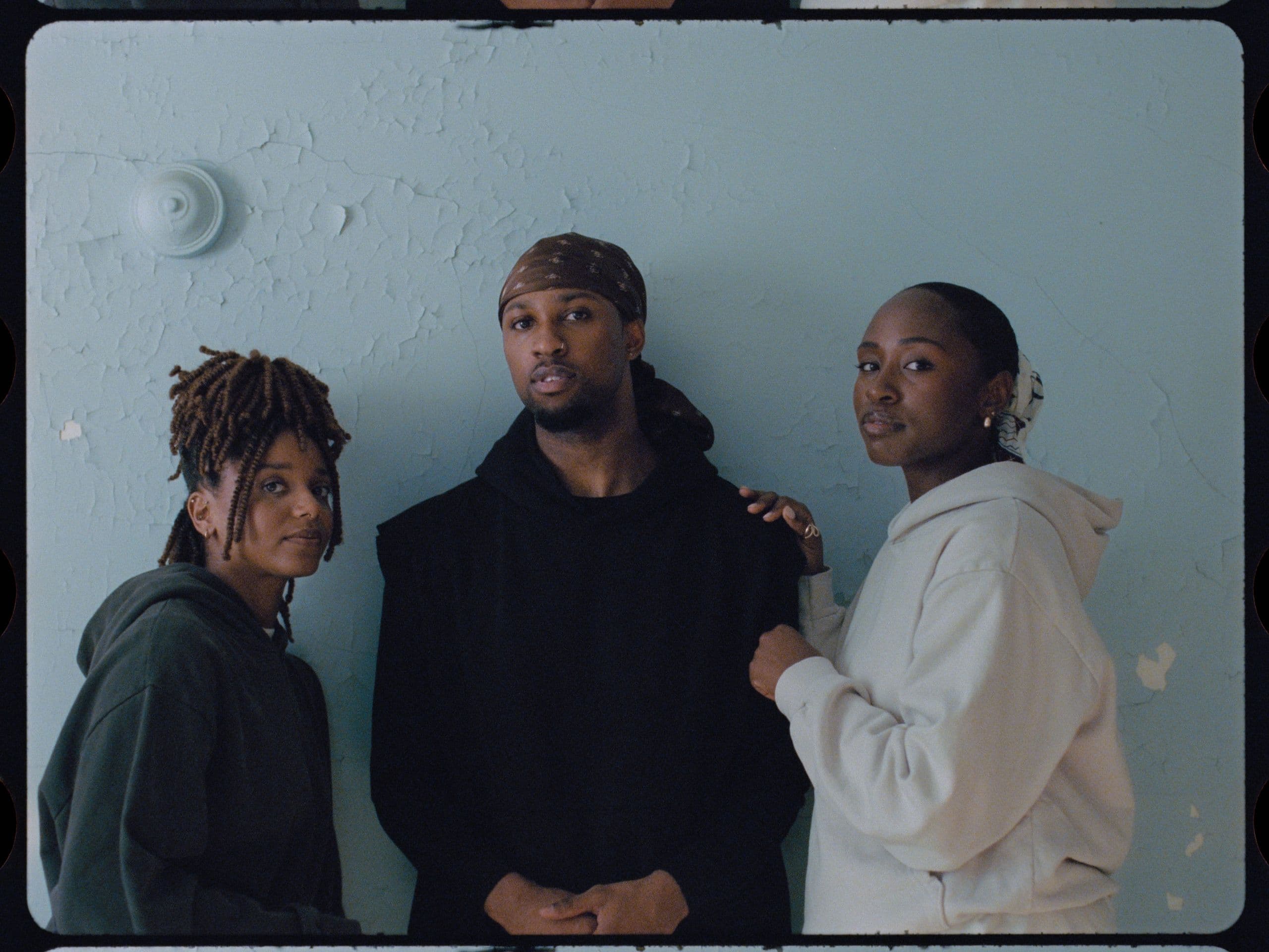Three dancers stand shoulder to shoulder against a pale blue wall with peeling paint, looking directly at the camera in muted hoodies and sweatshirts.
