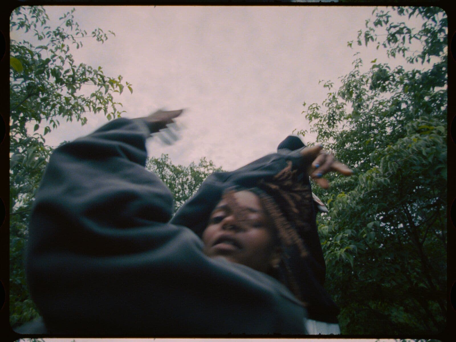 A candid, motion-blurred photo taken from a low angle looking up, showing a person mid-gesture with an arm raised, framed by tree branches against an overcast sky, with visible film-frame edges suggesting an analog 35mm still.