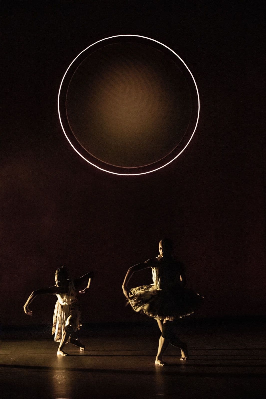 Two dancers performing under a large glowing ring suspended against a dark stage background.