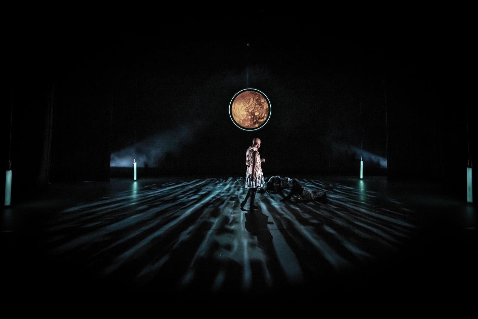 Wide view of a dark stage featuring a central dancer standing under a glowing ring, with water-like light reflections on the floor.