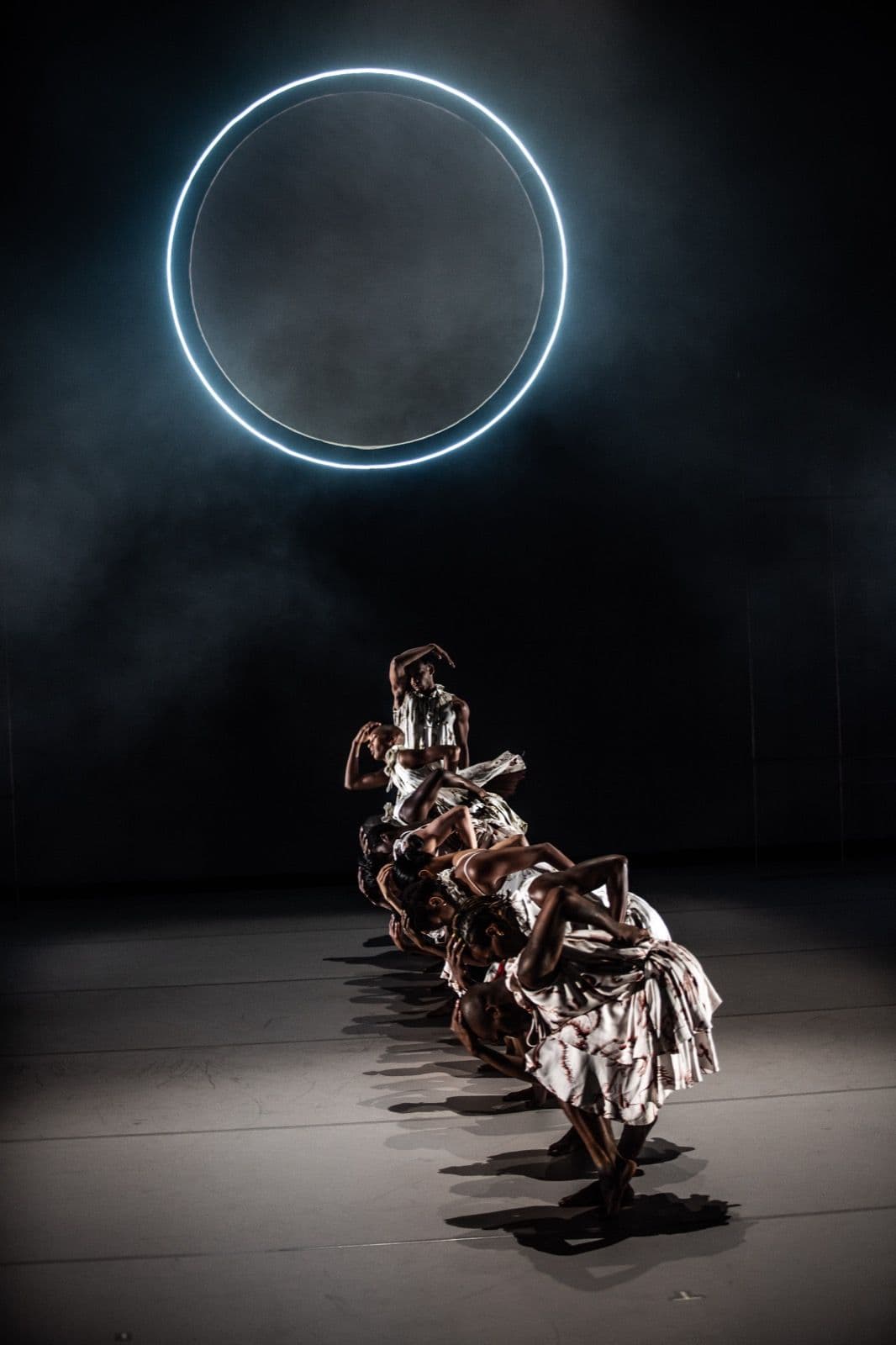 A line of dancers in matching patterned dresses crouching forward in unison beneath a large illuminated ring.