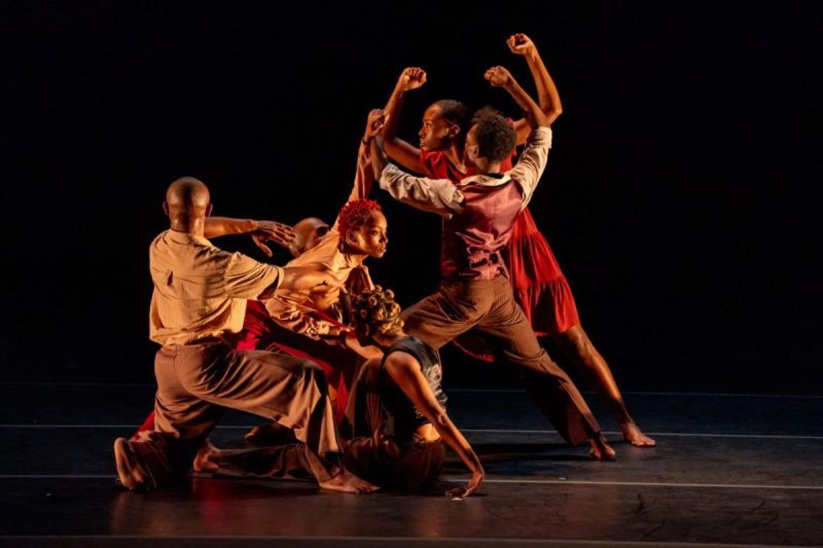 A group of contemporary dancers in warm-toned earth clothing form an expressive, clustered pose under warm stage lighting.