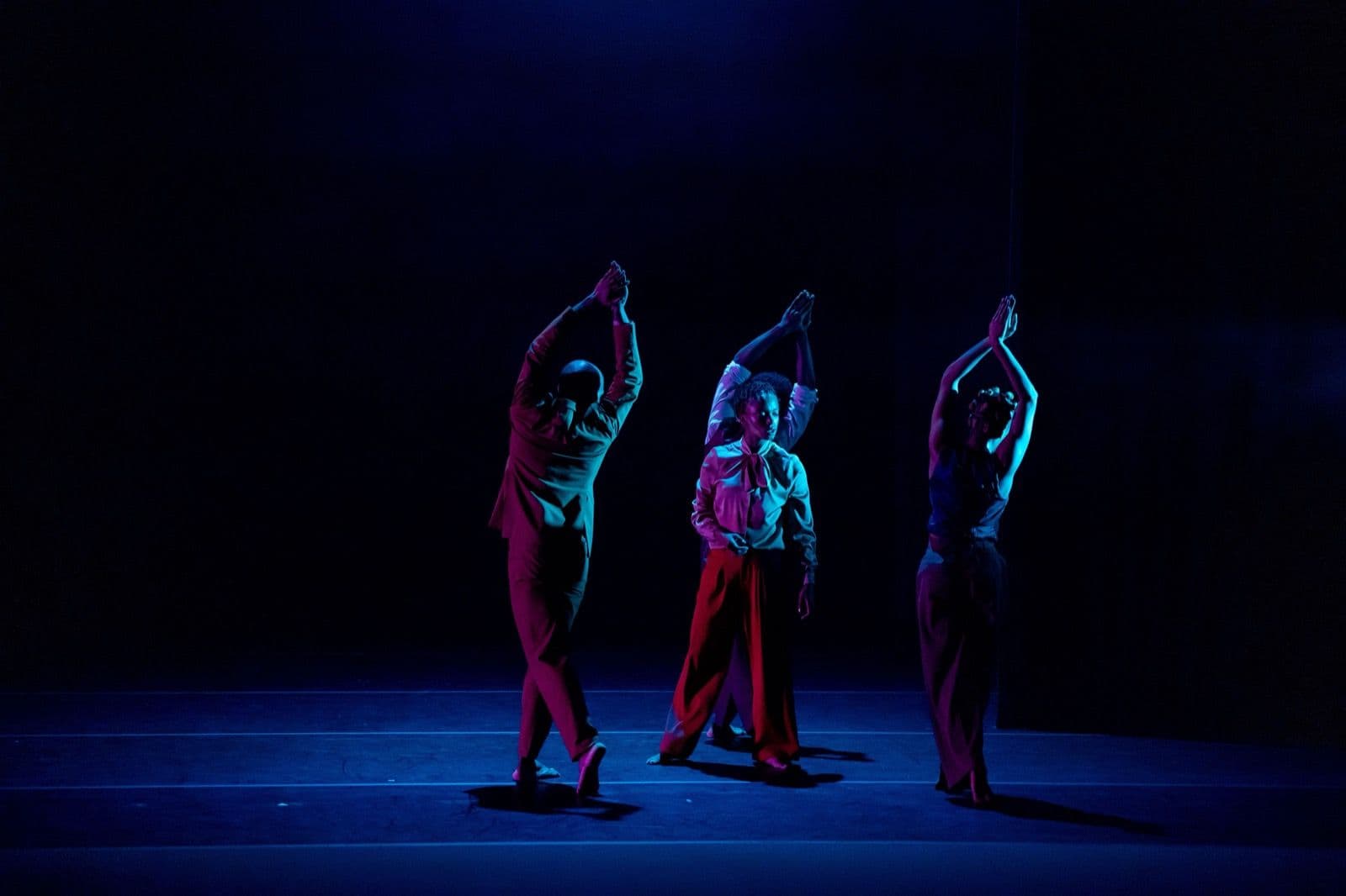 Four dancers perform under dramatic blue and purple stage lighting, holding overhead arm positions on a dark stage.