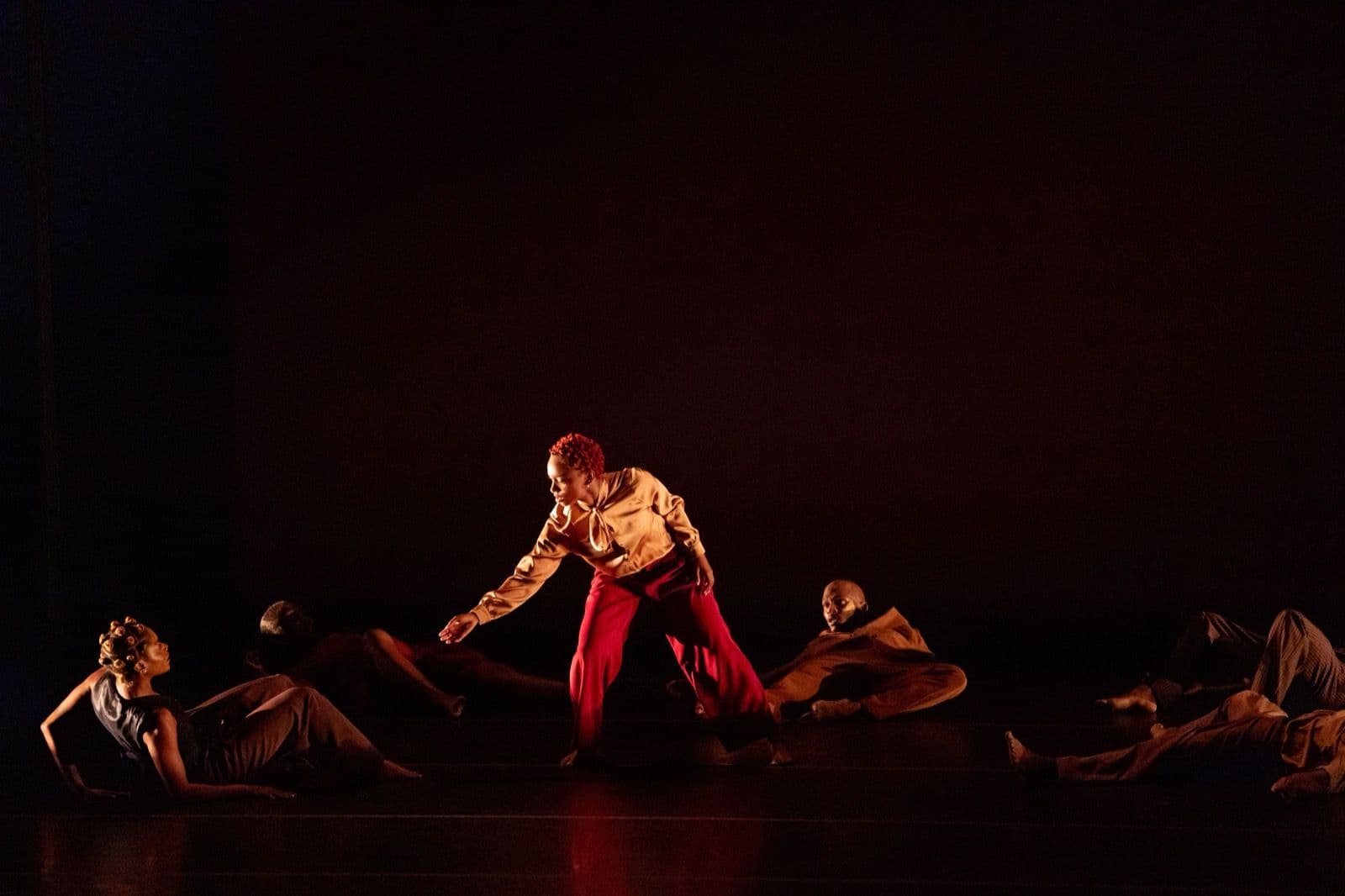 A soloist in red pants stands and reaches toward fellow dancers resting on the floor during a dark stage performance.