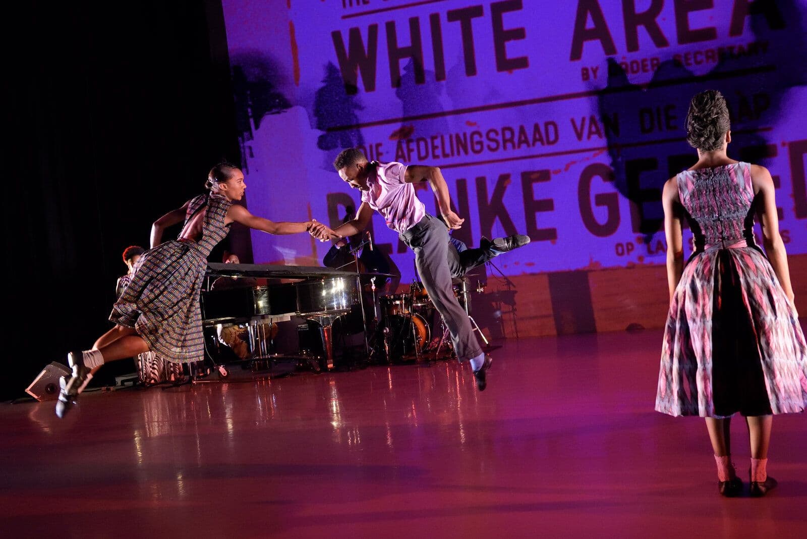 Three dancers in vintage-style patterned clothing move together near a grand piano and live band, one in a full skirt reaching toward two partners in a low lunge, lit in blue stage light with bold projected text overhead.
