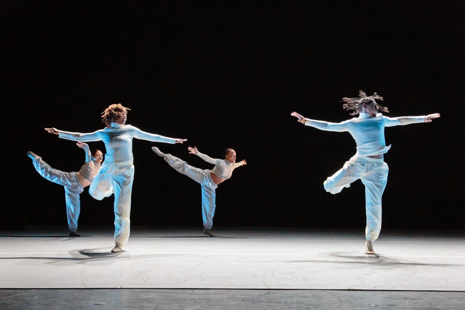 Four dancers from A.I.M by Kyle Abraham leap and extend their limbs across a dark stage in a synchronized contemporary dance performance, wearing matching light outfits.