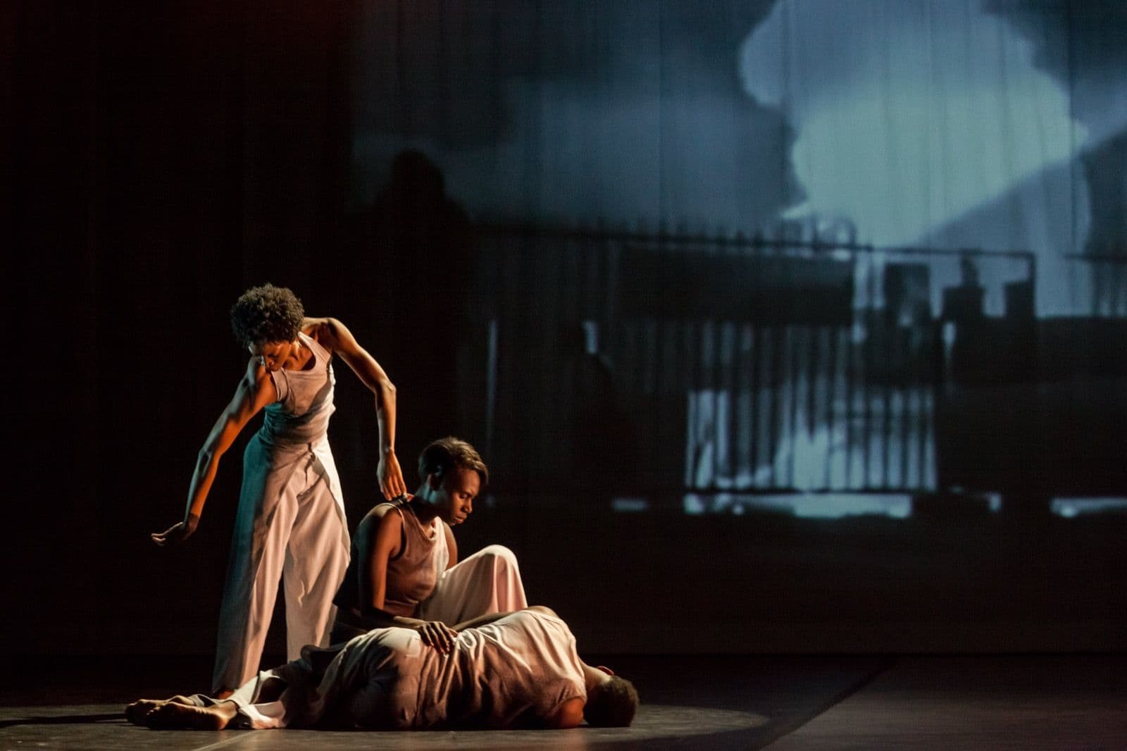Three dancers in light-colored clothing perform a solemn, grounded pose on a dimly lit stage in "Absent Matter."