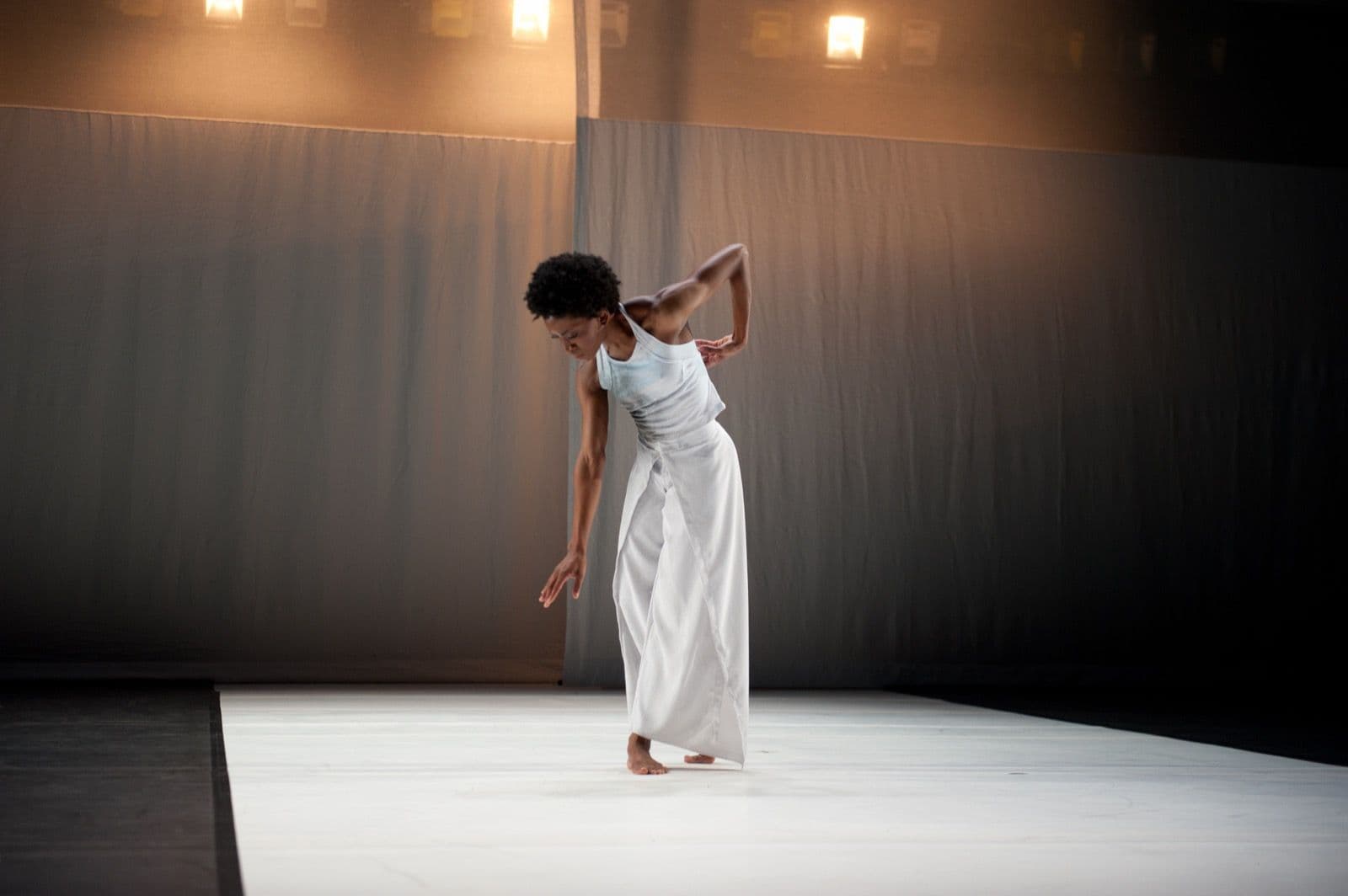 Solo dancer Tamisha A. Guy bows slightly and reaches one arm downward during a contemporary dance performance on a white floor.