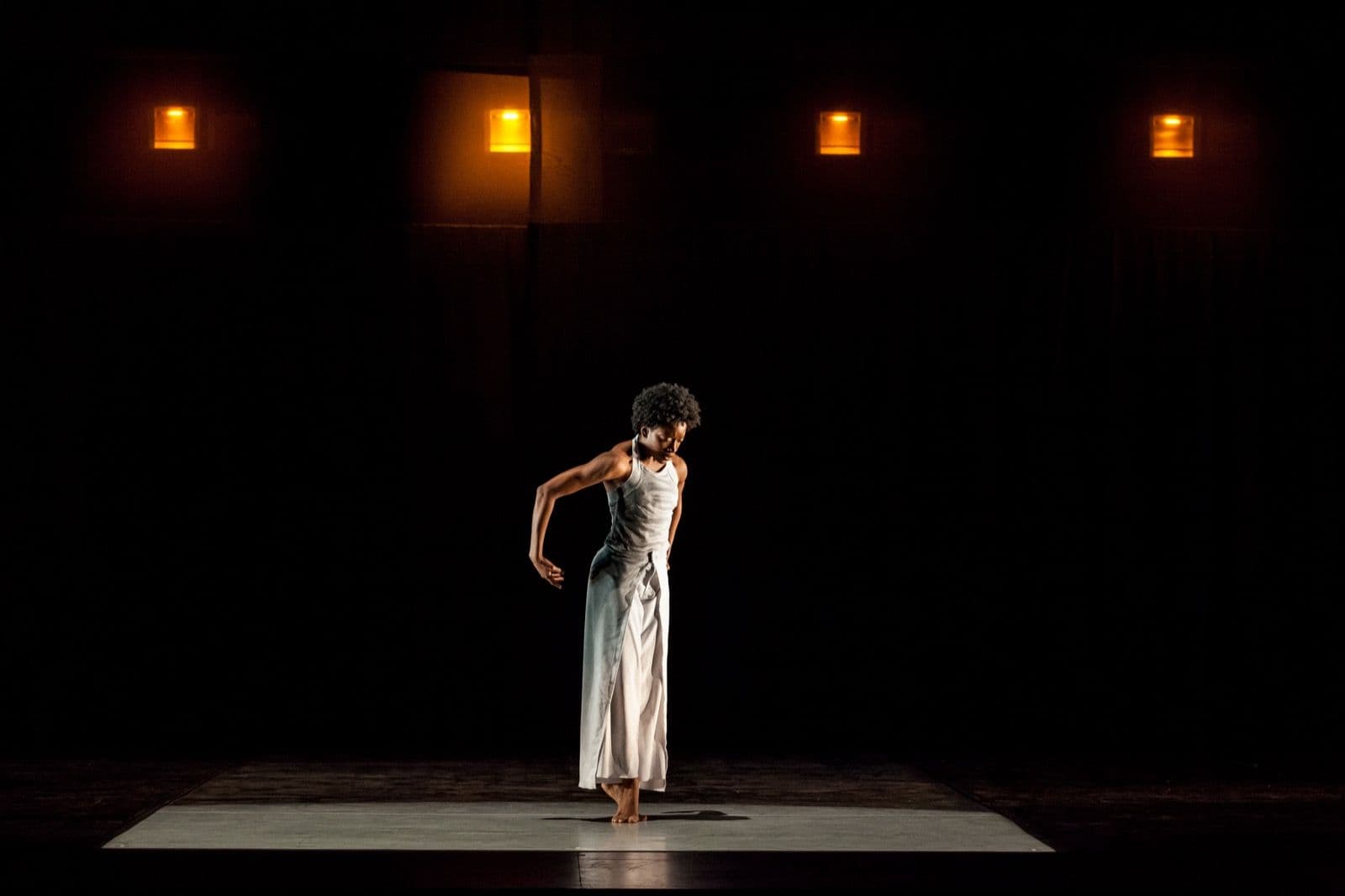 Dancer Tamisha A. Guy stands centered under overhead lights with her head bowed and hands resting near her hips.