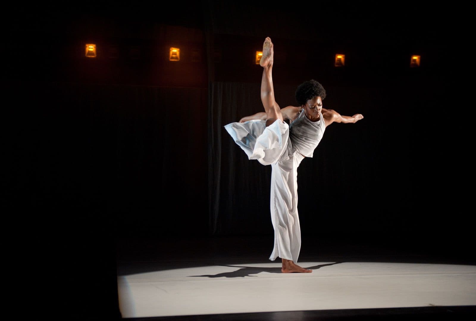 Dancer Tamisha A. Guy executes a high vertical leg extension with wide arms under stage spotlights.