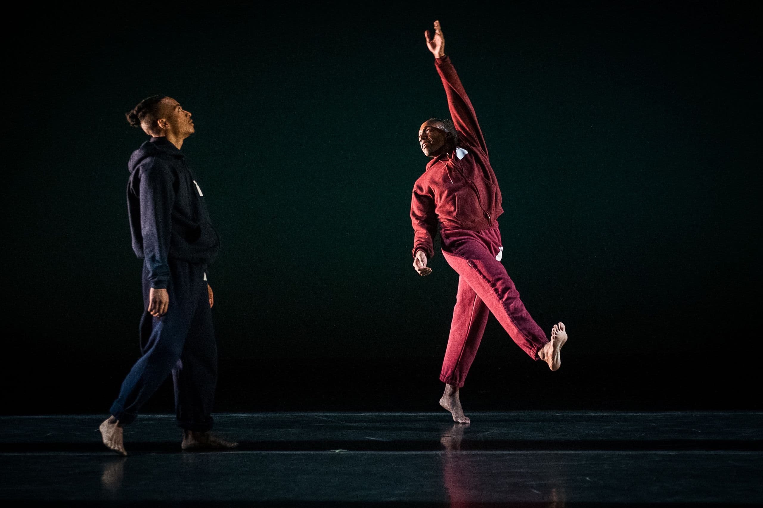 Two barefoot contemporary dancers performing on a dark stage; one stands in a dark blue sweatsuit looking upward as the other in a red sweatsuit reaches one arm high and extends a leg forward.
