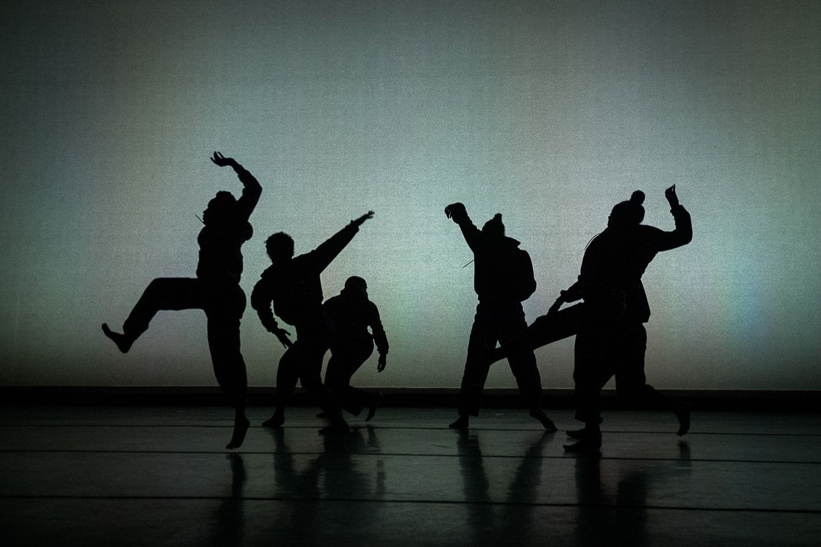 Five dancers stand in silhouette against a dimly lit background, frozen mid-movement during a performance of "Big Rings."