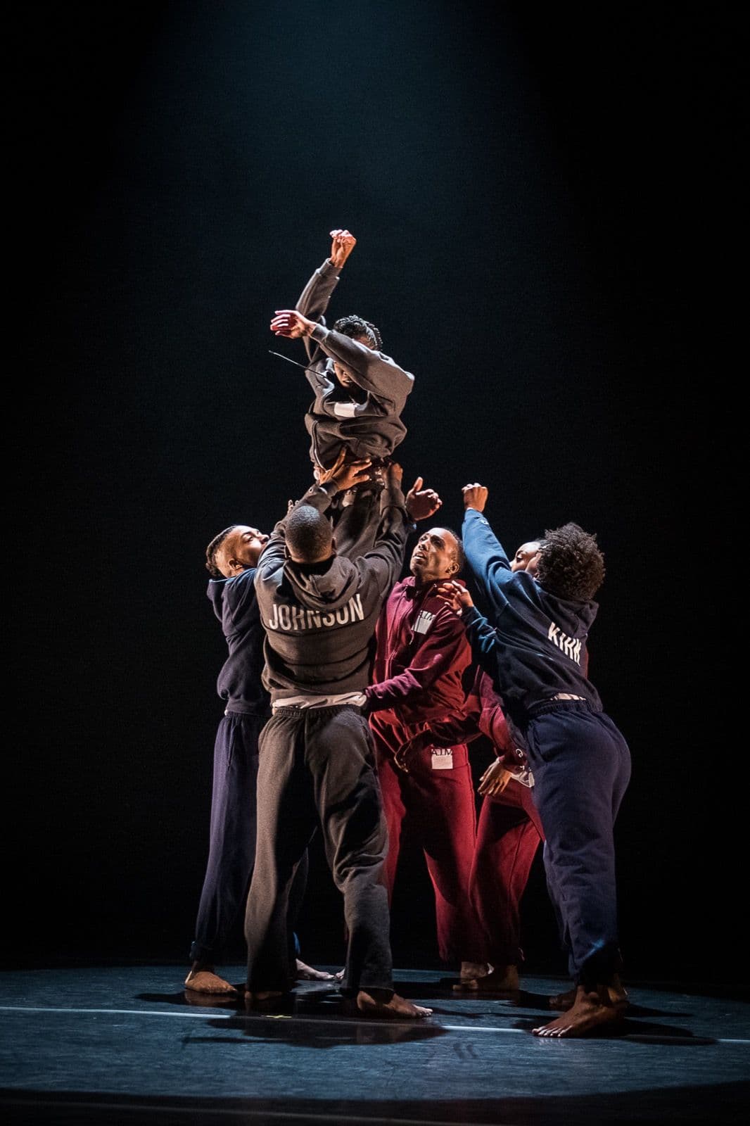 Dancers in sweatpants and hoodies gather under a spotlight to lift a fellow dancer high into the air.