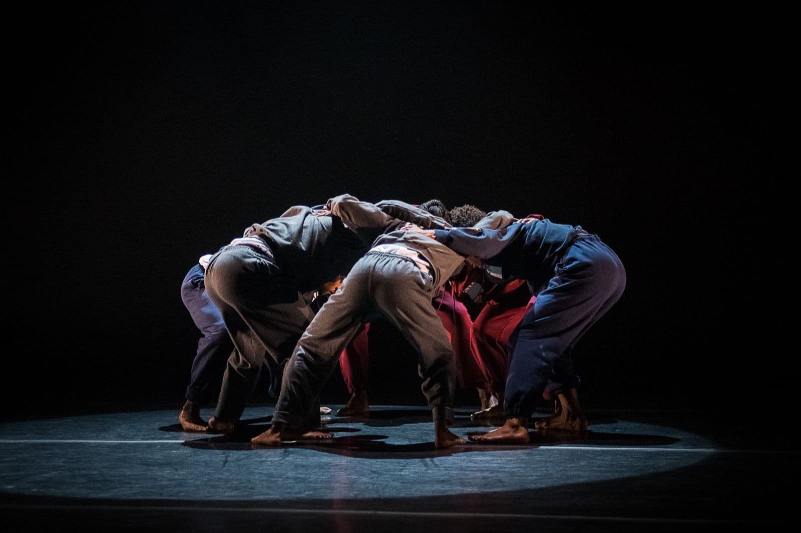 A group of dancers clad in tracksuits hunch forward in a tight huddle beneath a single pool of light.