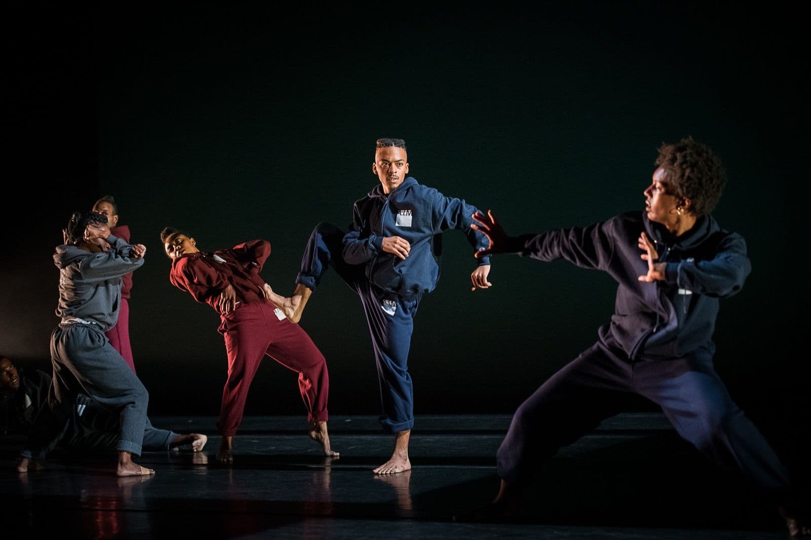 Dancers wearing hoodies strike sharp, energetic athletic poses across a dark stage in "Big Rings."