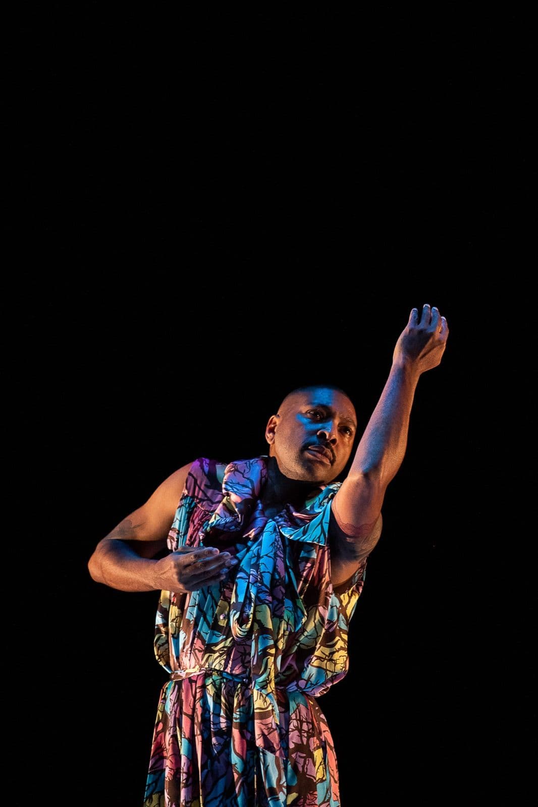 Choreographer Kyle Abraham performs solo in a colorful, patterned outfit on a dark stage, extending one arm gracefully upward.