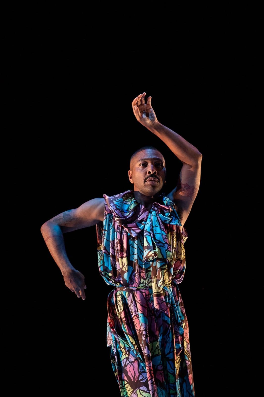 Kyle Abraham dances solo in a vibrant patterned jumpsuit, lifting one bent arm above his head.