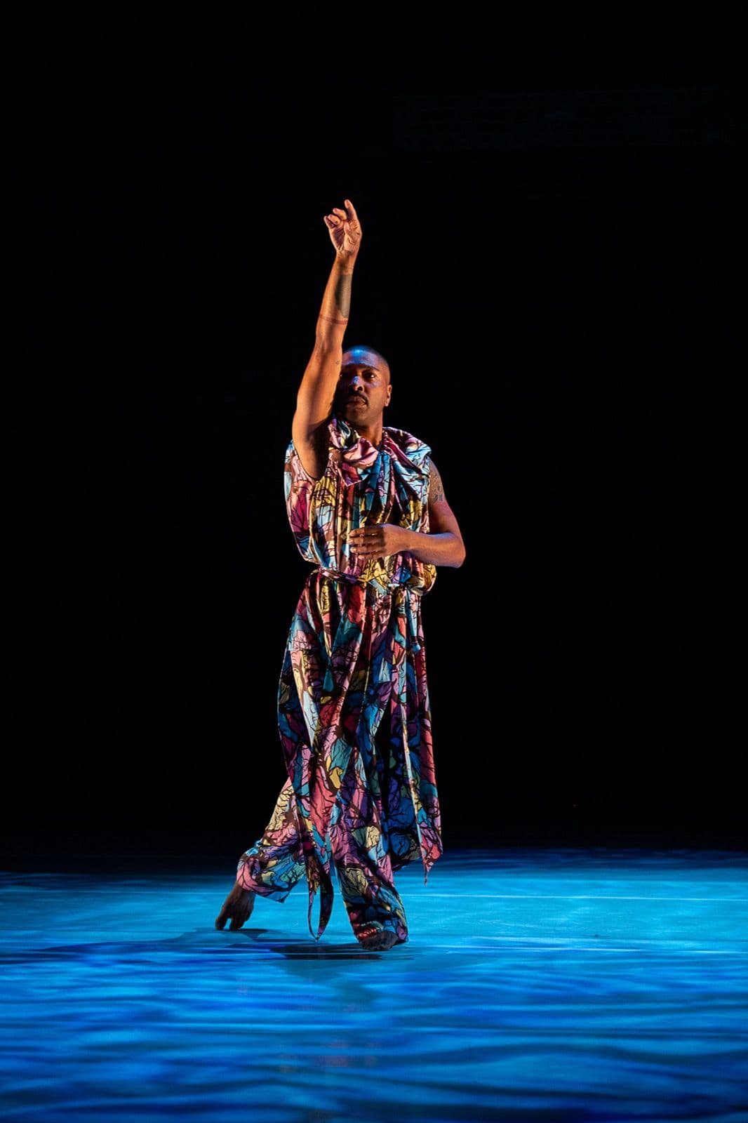 Solo dancer Kyle Abraham stands on a stage floor illuminated by blue light, extending his arm high into the air.
