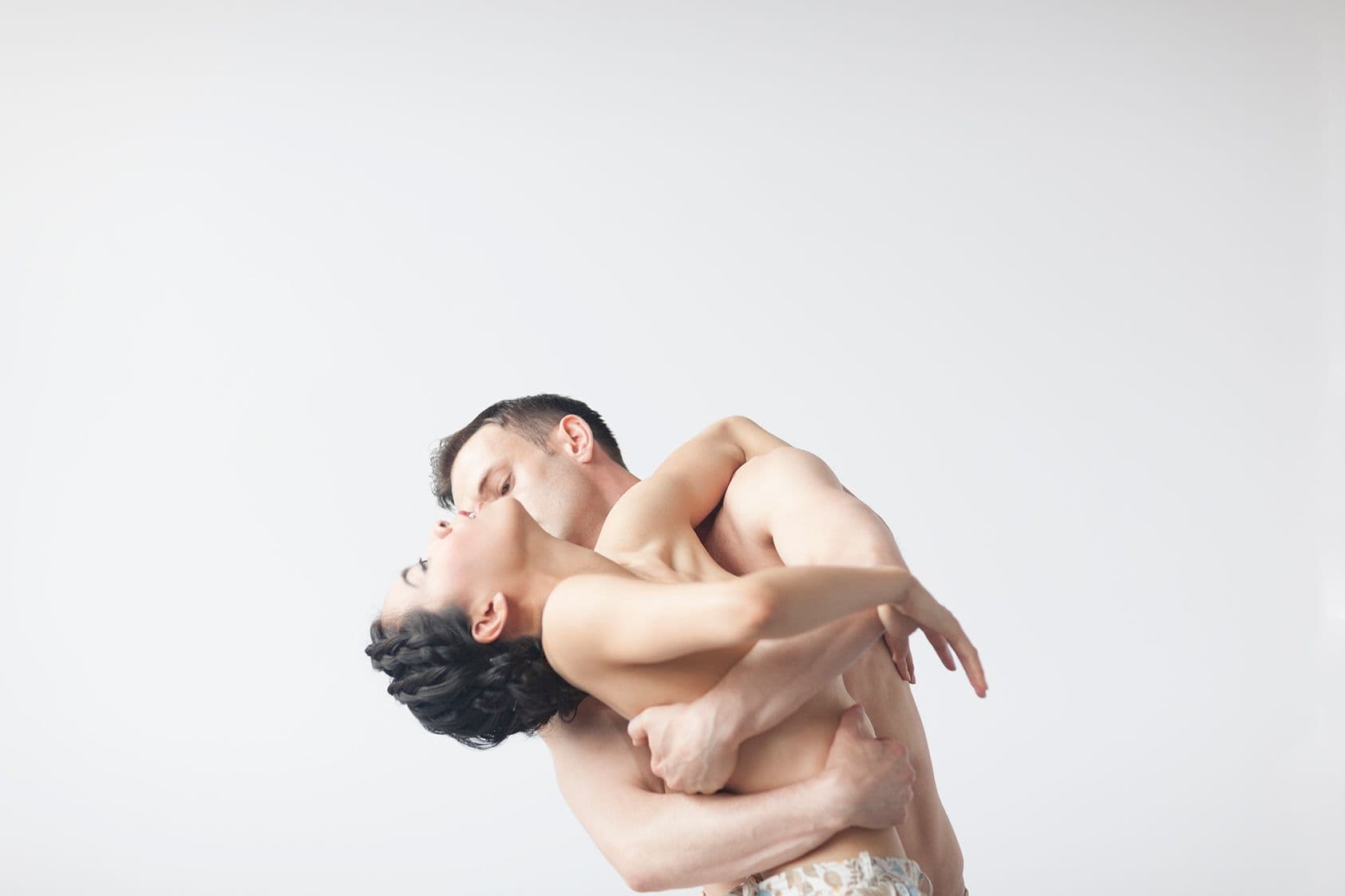 Two dancers intertwined, torsos bare, arms wrapped tightly around each other with faces close together in profile, against a plain white studio background.
