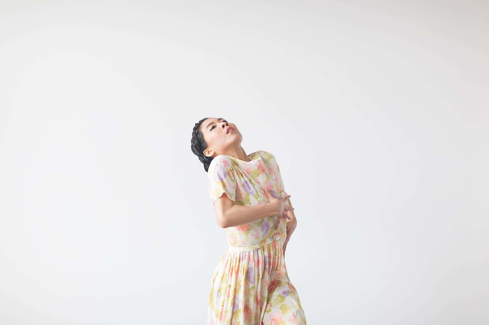 A dancer in a pastel floral dress arches backward with her head tilted up and one hand resting on her waist, set against a plain white studio background.