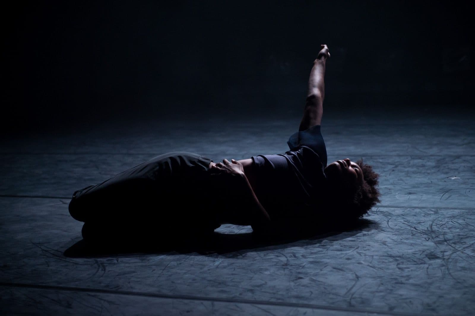 Solo dancer Catherine Ellis Kirk lies on her side on a dimly lit stage with one arm extended overhead.