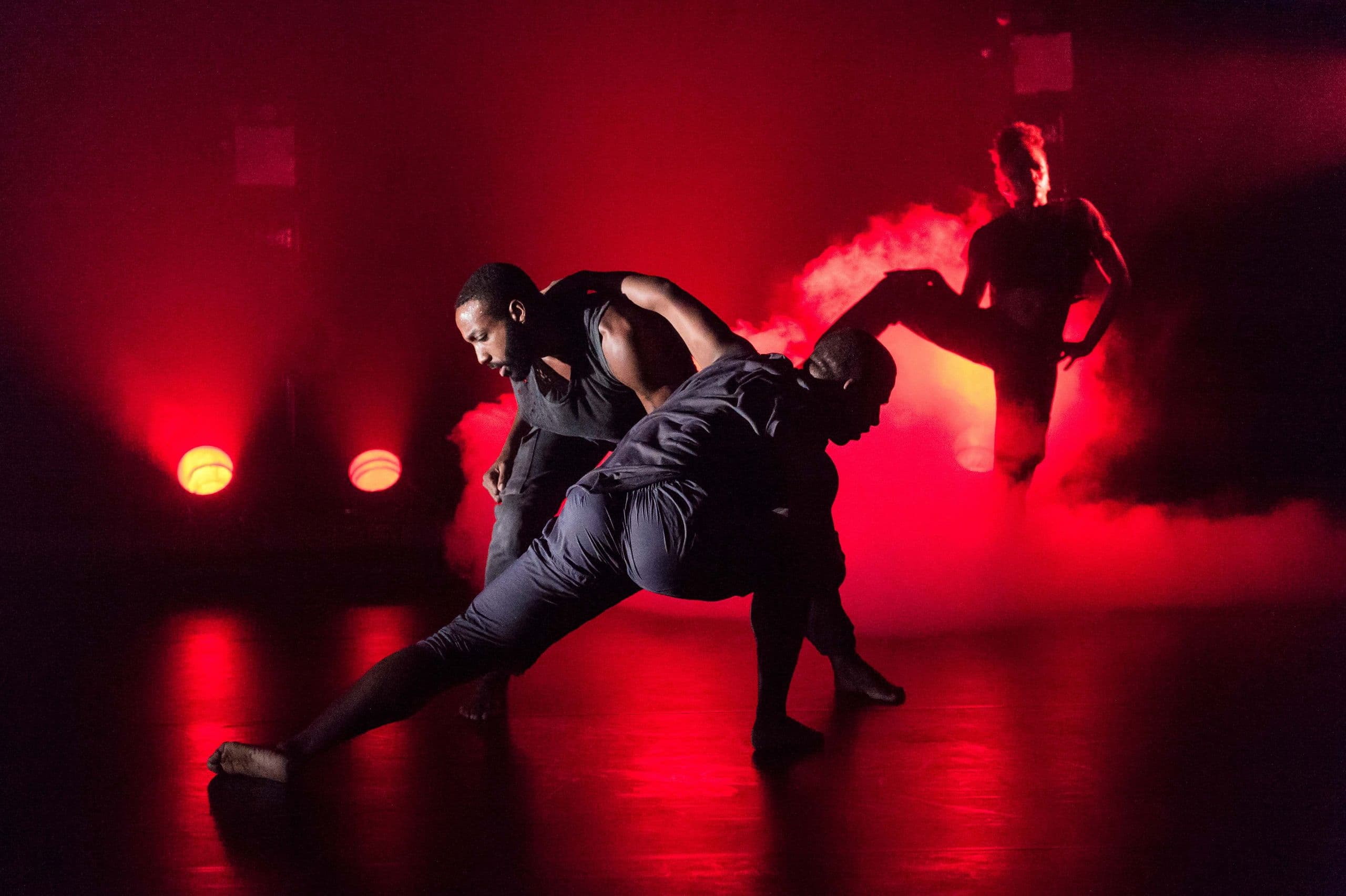 Three contemporary dancers performing on a stage bathed in intense red lighting and atmospheric smoke; two male dancers lean in close near the center while a third dancer stands silhouetted in the background.