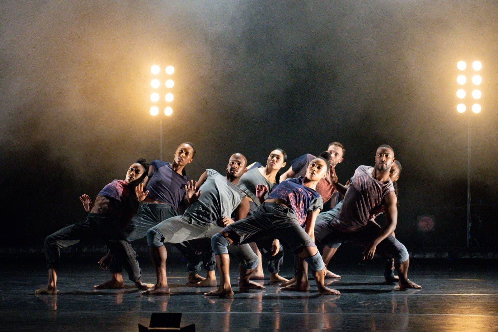 An ensemble of A.I.M by Kyle Abraham dancers hit a synchronized low stance on a dimly lit stage flanked by vertical light rigs.