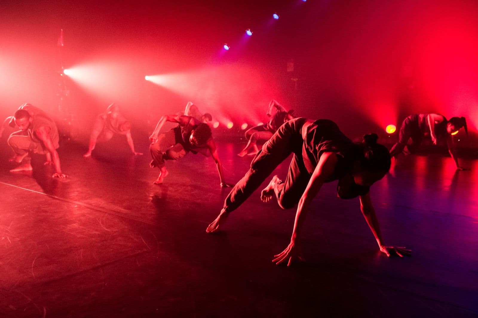 Dancers crouch and shift low across a stage bathed in intense red lighting and atmospheric haze.