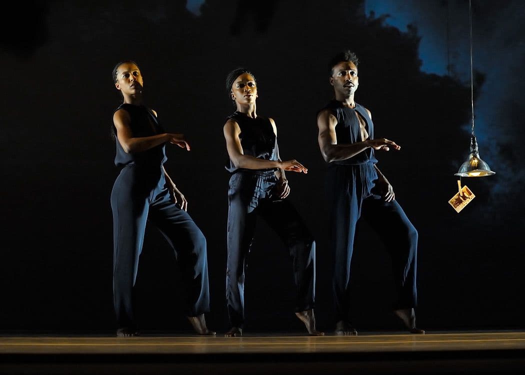 Three dancers in matching dark outfits perform in unison under a low-hanging pendant light on a dimly lit stage.