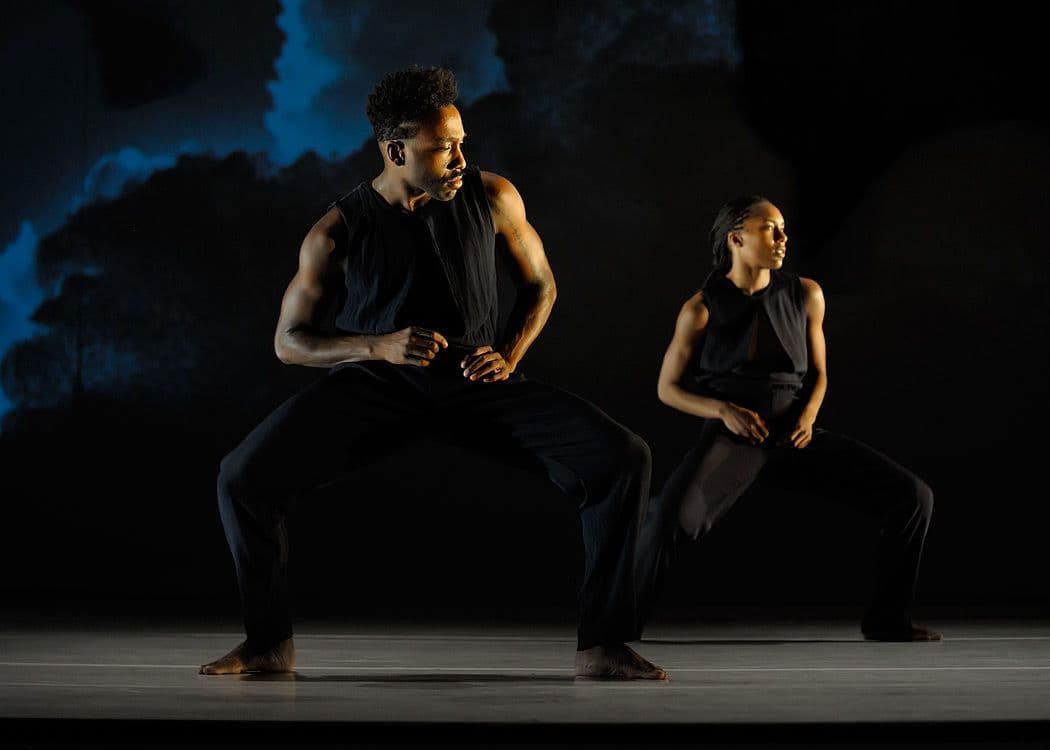 Two dancers in dark sleeveless outfits perform in wide, grounded stances against a moody blue-lit backdrop.