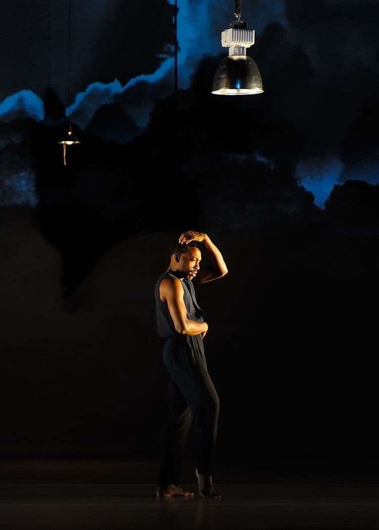 A male dancer in a sleeveless dark jumpsuit stands under a single spotlight with one hand raised to his head.