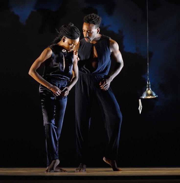 A male dancer gently rests his hand on a female dancer's shoulder beside a hanging lamp in a dark performance space.
