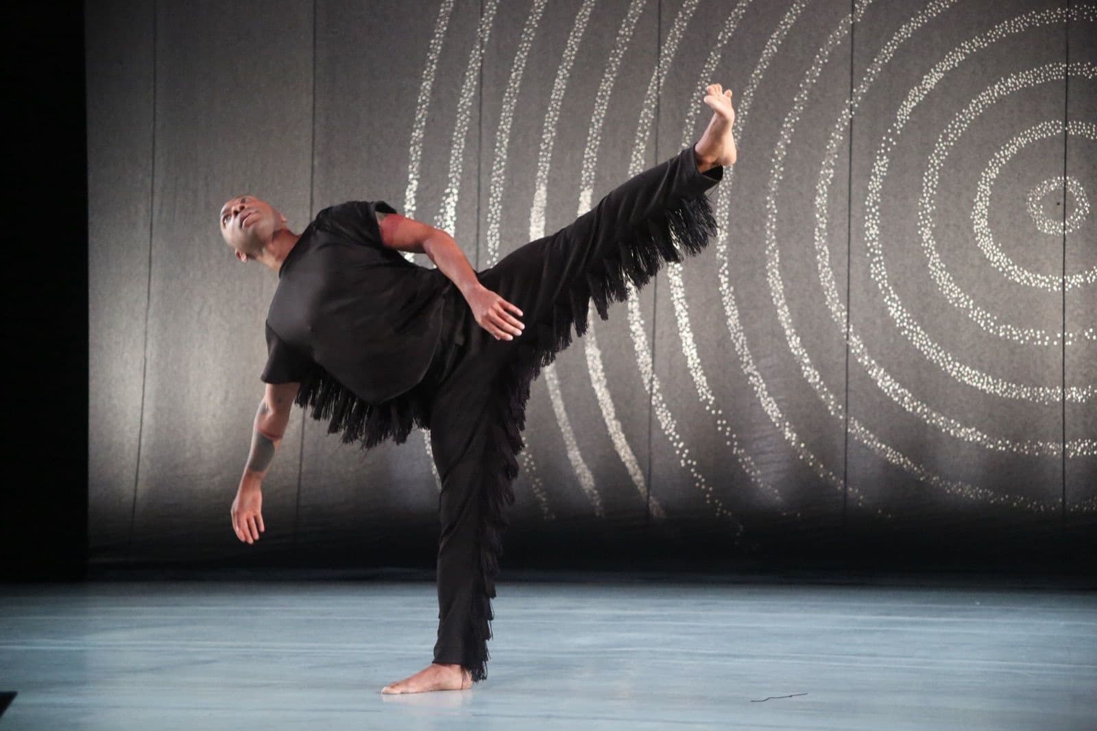 Contemporary dancer Kyle Abraham performs on stage in a black fringed outfit, lifting one leg high in an extension against a backdrop of glowing concentric circles.