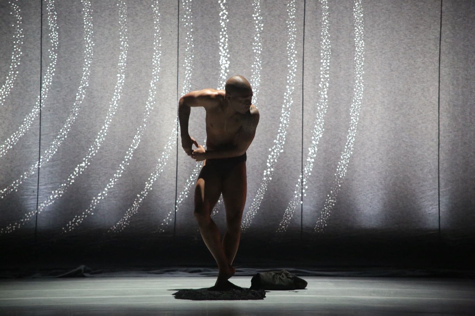 Bare-chested dancer Kyle Abraham strikes a fluid, bowed posture on stage against a backdrop of curved, illuminated dot patterns.