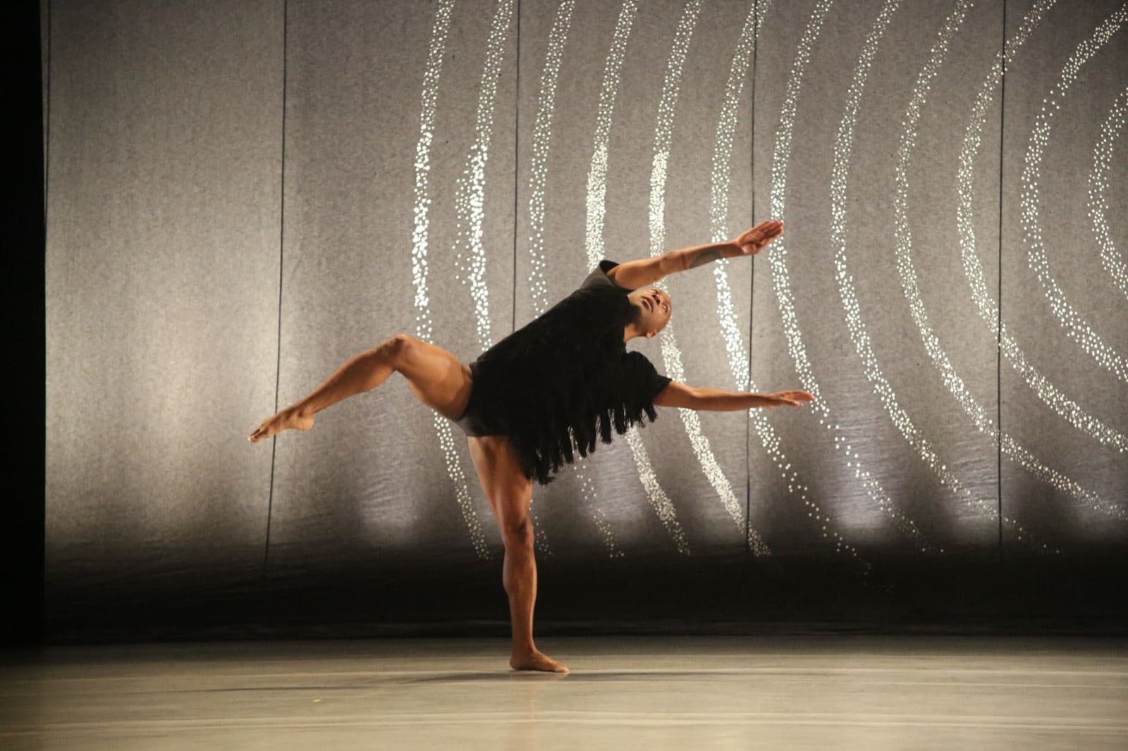 Dancer Kyle Abraham balances dynamically on one leg in a black fringed top, extending his arms and opposite leg against a patterned stage backdrop.