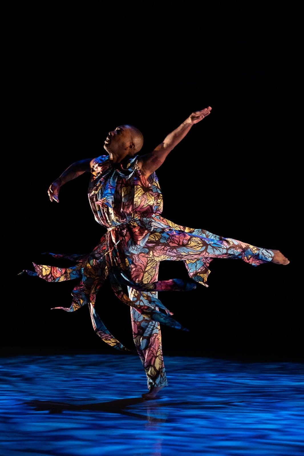 Kyle Abraham performs an expressive solo leap across a blue-lit stage, wearing a flowing, colorful patterned costume.