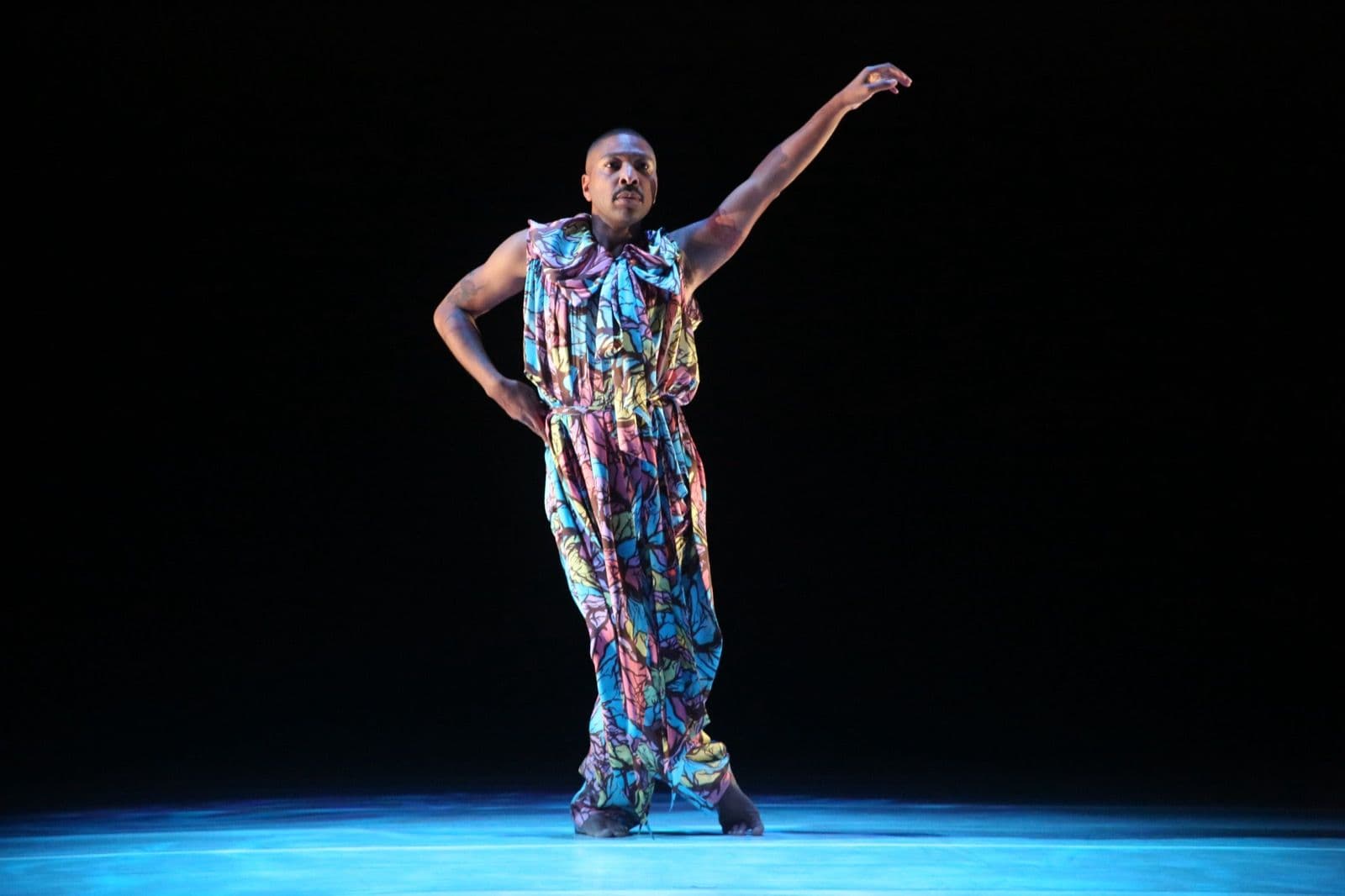 Kyle Abraham stands on a blue-lit stage in a colorful garment, extending one arm outward in a fluid gesture.