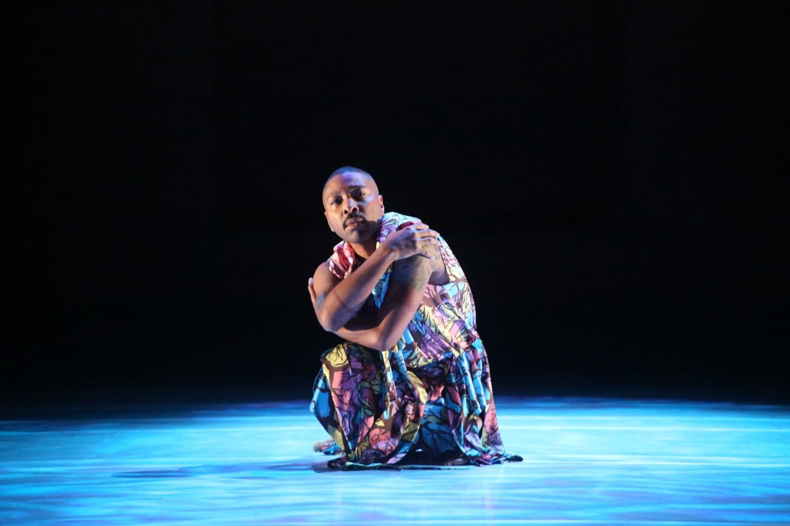 Kyle Abraham crouches low on stage, wrapping his arms around himself while gazing toward the audience.