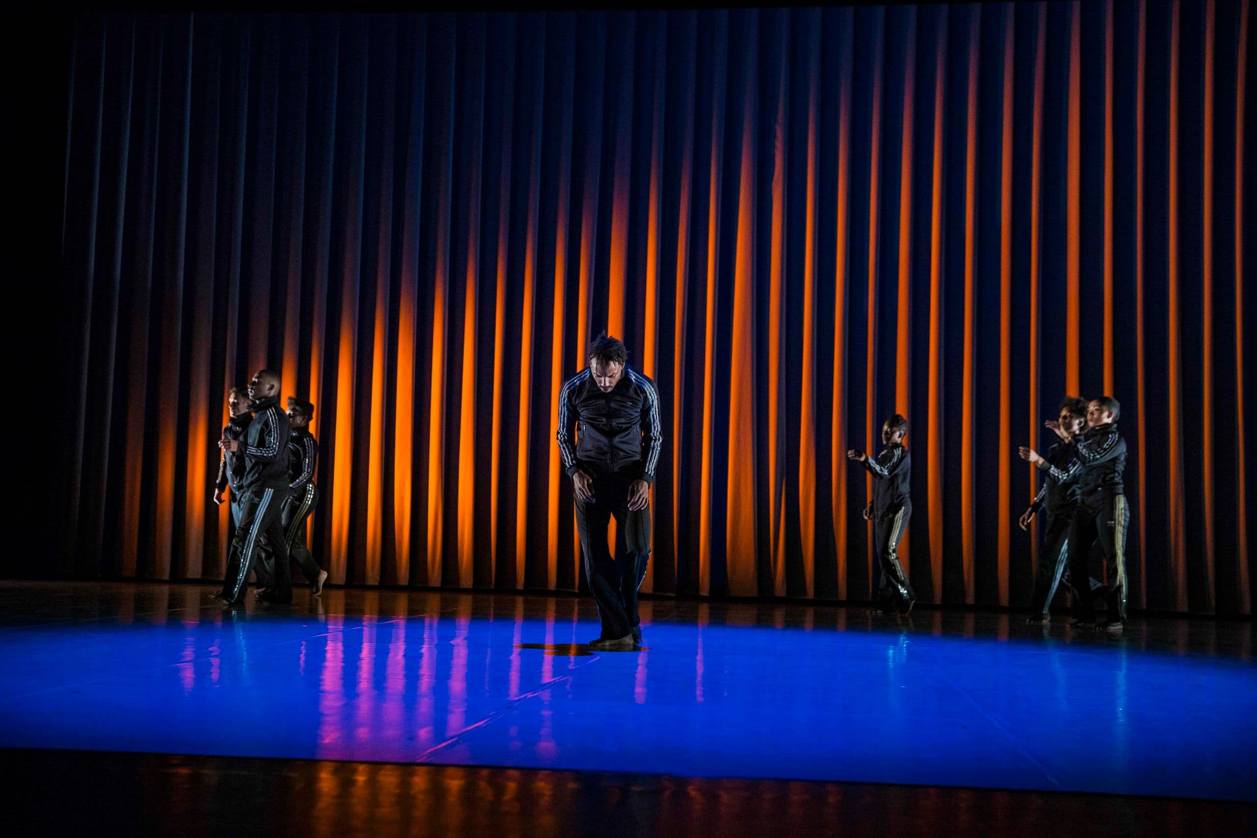 A group of dancers in matching dark tracksuits performing on a stage with a blue-lit floor and a striped orange-and-black backdrop, featuring one soloist bowed at center stage.