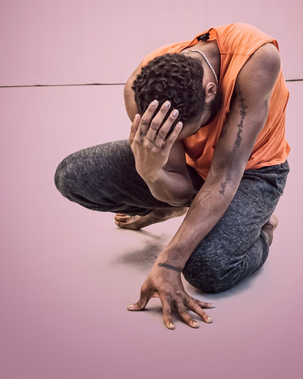 Dancer Jae Neal crouched low in an orange sleeveless top with one hand over face and one on the floor.