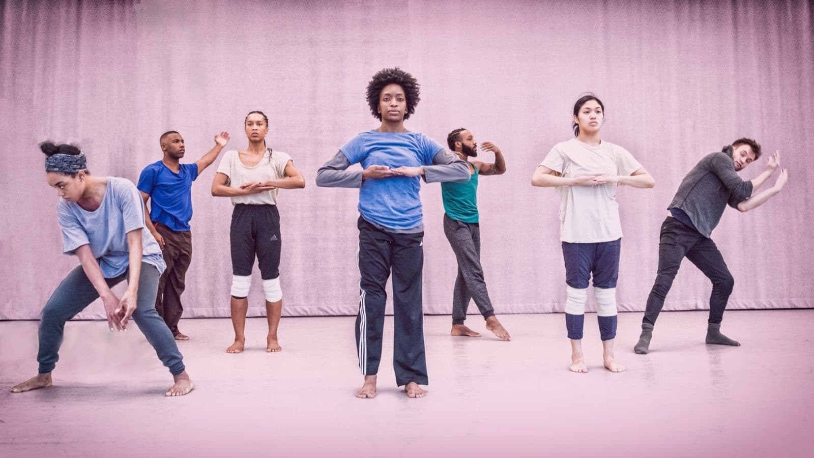 Group of dancers spread out on stage performing choreographed movements in front of a light curtain.