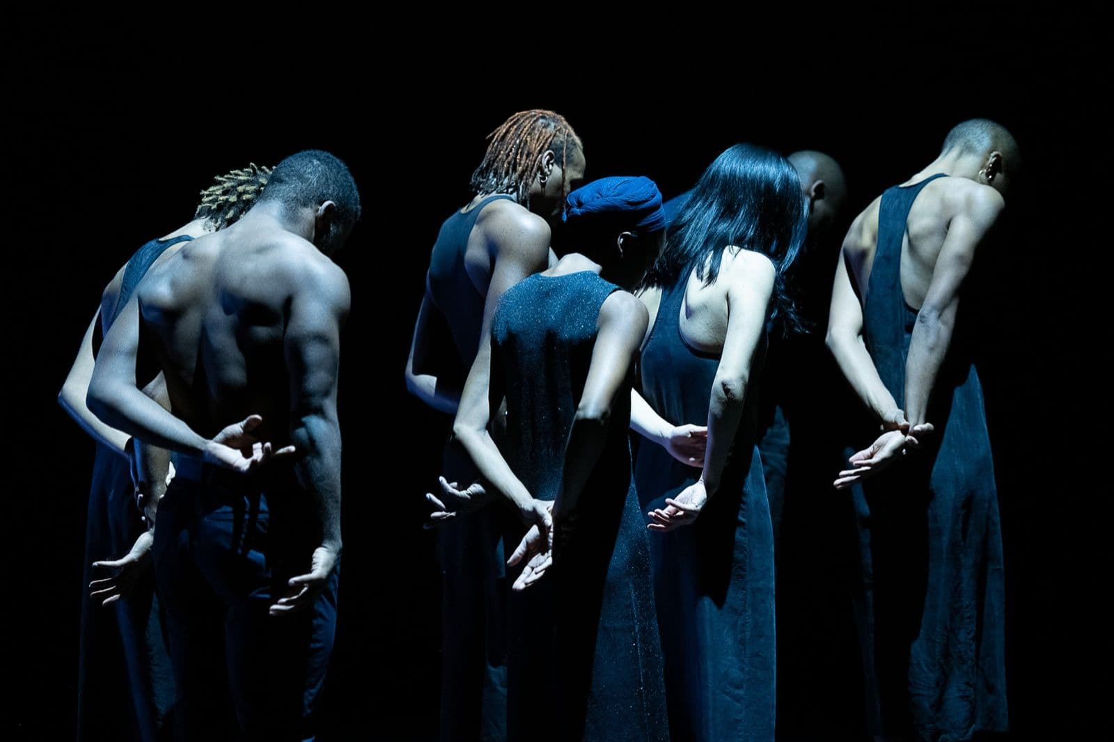 A line of dancers in dark clothing standing with their backs to the camera under cool blue lighting, hands clasped behind them.
