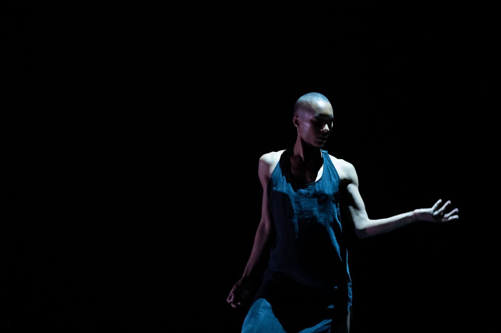 A solo dancer with short hair in a dark tank top performing under dramatic spotlight against a black background.