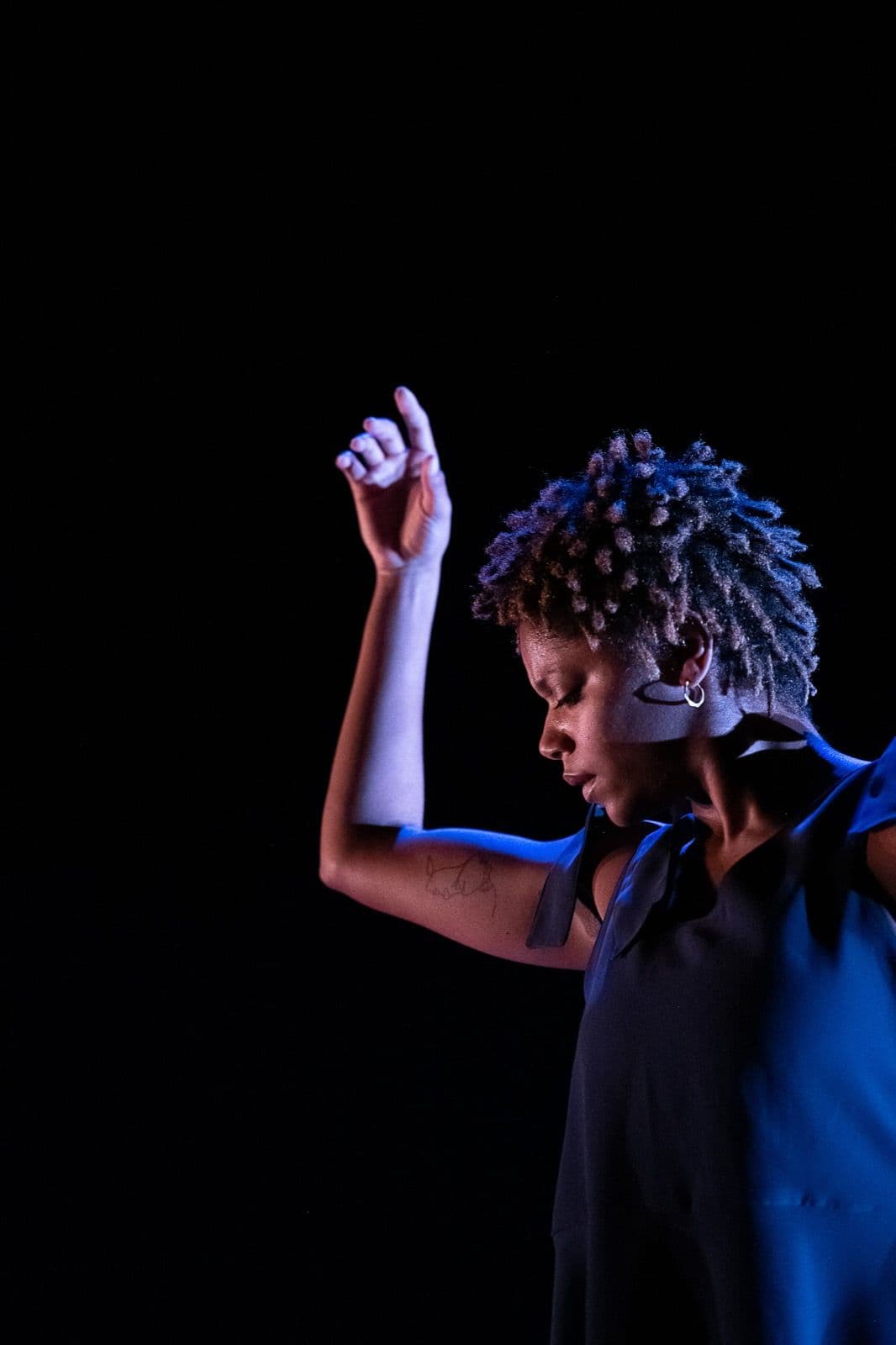 A close-up side profile of a dancer with short textured hair extending one arm upward in soft blue lighting.