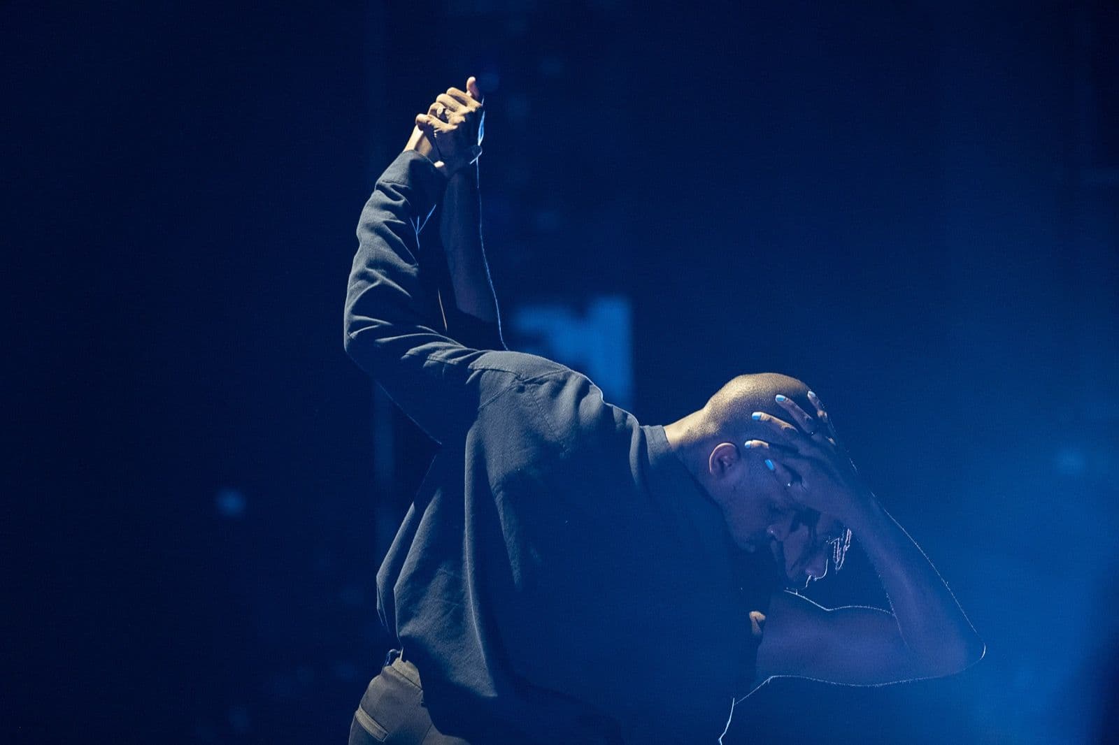 Two male dancers intertwined under blue light, one holding their hands overhead while the other rests a hand on his head.
